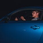 Młody mężczyzna w okularach prowadzi niebieski sportowy samochód nocą